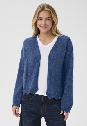 Un cardigan en tricot bleu présente une coupe ample, un ourlet festonné et un tissu texturé. Porté par-dessus un t-shirt blanc à col en V.