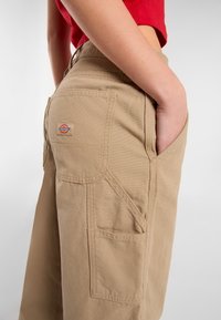 Beige cargobroek met grote zijzakken, versterkte stiksels en een klein merkembleem. De stof lijkt duurzaam met een gladde textuur.