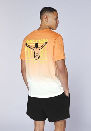 Mann trägt ein T-Shirt mit orange-weißem Farbverlauf und schwarzer Grafik eines Tauchers von hinten sowie dem Text „SINCE 1982 CHI SEE Active Wear“, kombiniert mit schwarzen Shorts.