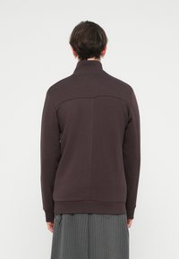Rjava zip-up sweatshirt z visokim ovratnikom, rebrastimi manšetami in teksturirano tkanino. Ima vodoravno šivanko čez zgornji del hrbta.