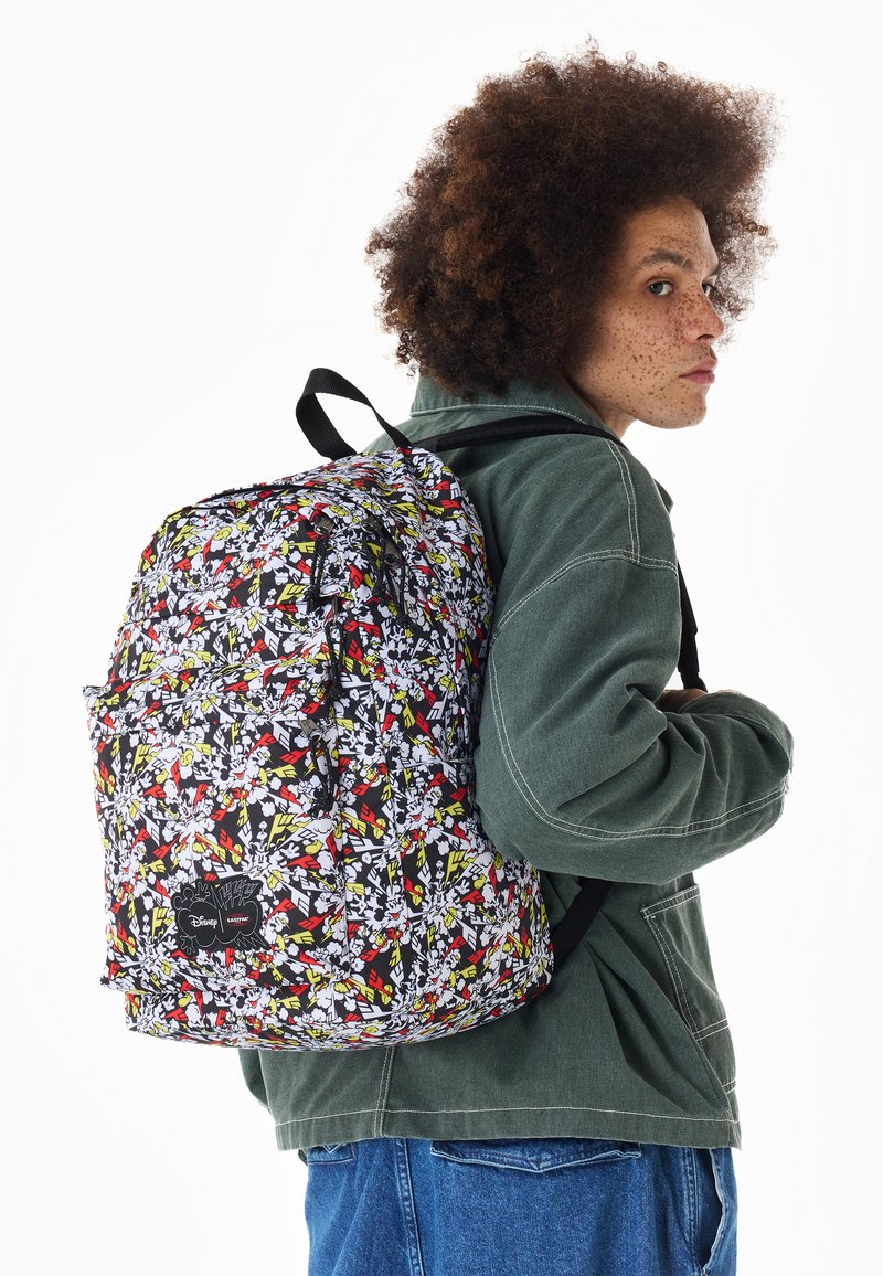 Person mit lockigem Haar, die eine grüne Jacke und blaue Jeans trägt, trägt einen großen bunten Rucksack über einer Schulter.