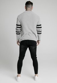 Man med nacktattoo som bär en grå tröja med svarta ränder, svarta jeans och vita sneakers, stående vänd mot en plain grå bakgrund.