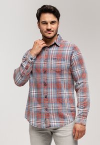 Mężczyzna poprawiający kołnierzyk, ubrany w niebieską, białą i czerwoną koszulę w kratę z kieszenią na piersi oraz jasnoszare spodnie, na tle jednolitego tła.