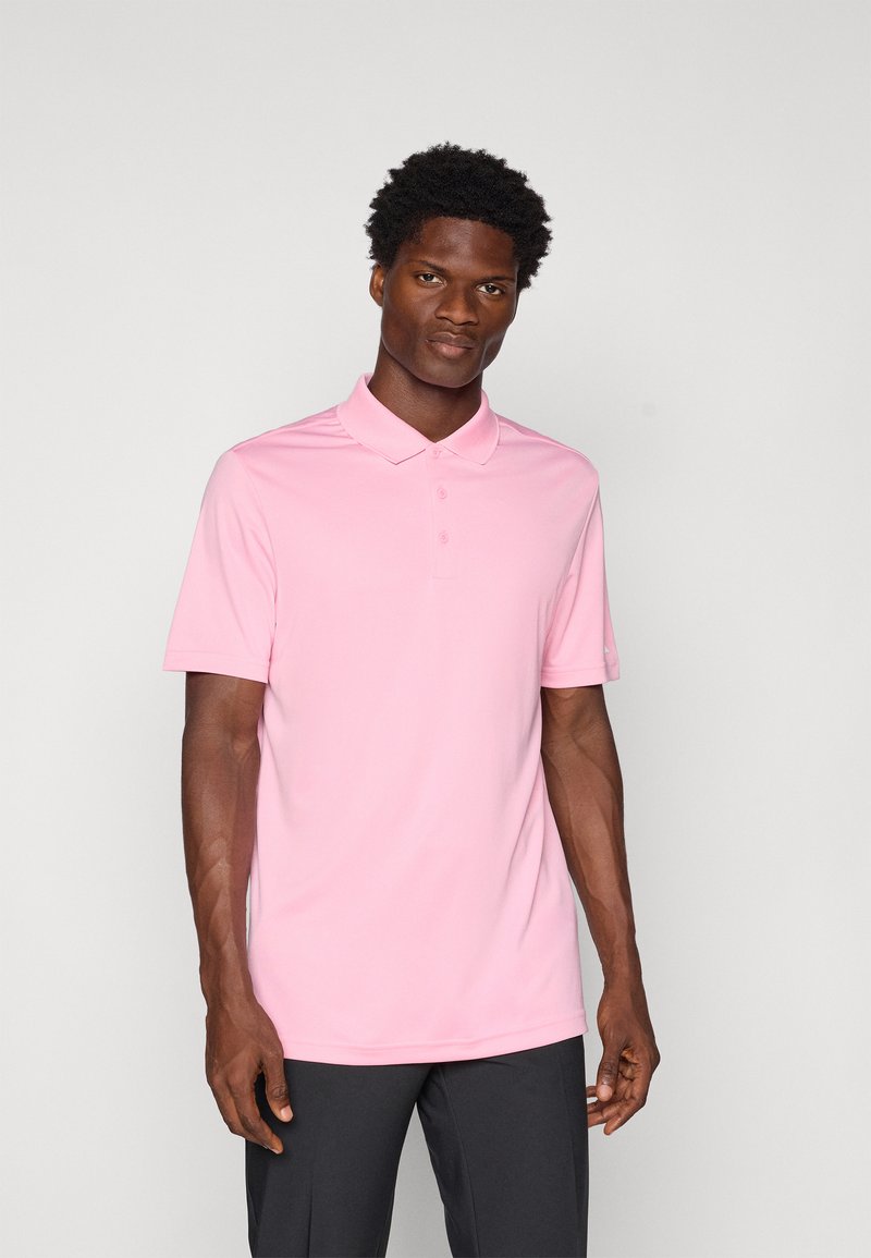 Πόλο μπλούζα σε ανοιχτό ροζ χρώμα, κοντά μανίκια, με κουμπωτή πατιλέτα, λεία υφή, φορεμένη με σκούρους παντελόνες, κεντραρισμένη σε ουδέτερο φόντο.