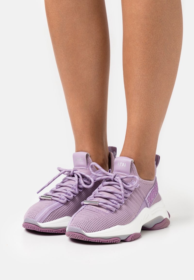 Jambes portant des baskets en mesh lilas avec des semelles épaisses blanches et des lacets, debout sur une surface blanche.