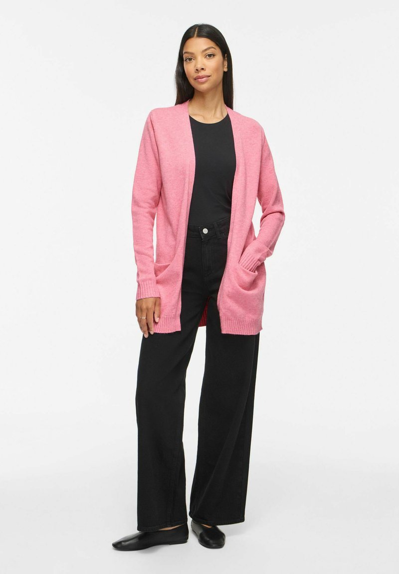 Femme portant un cardigan rose ouvert devant, une chemise noire, un pantalon noir à jambes larges et des ballerines noires, debout avec une main dans la poche.