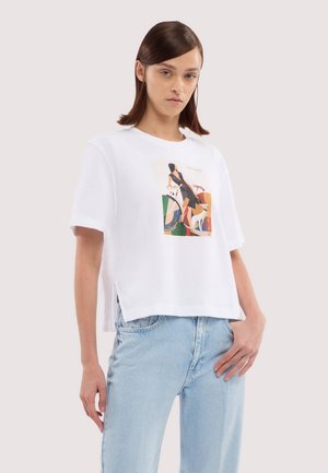 Kobieta o prostych brązowych włosach, ubrana w biały t-shirt z nadrukiem i jasnoniebieskie dżinsy, stoi na jednolitym jasnym tle.