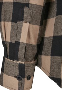 Plaid shirt in tan and black fabric, featuring a buttoned cuff, distinct check pattern, and neatly stitched seams. Textured surface.