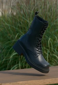 Svart läderankelhär boot med en tjock sula, featuring snören, en rundad tå och förstärkta sömmar. Klädd mot en naturlig bakgrund.