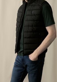 Czarna kamizelka puchowa z pikowanym wzorem, zapinana na zamek, z kieszeniami po bokach. Noszona na ciemnozieloną koszulkę i zestawiona z niebieskimi dżinsami.