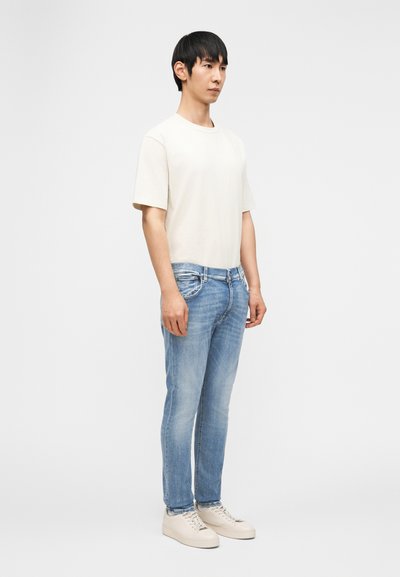 Νεαρός άντρας που φοράει ανοιχτό μπεζ μπλουζάκι, ανοιχτό μπλε τζιν και λευκά αθλητικά παπούτσια, στέκεται μπροστά σε λευκό φόντο.