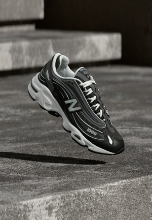 Baskets New Balance 1000 en noir et blanc avec des lacets blancs, suspendues au-dessus d'une surface en béton rugueuse avec des marches en pierre en arrière-plan.