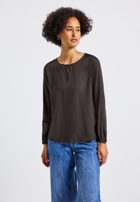 Donkerbruin blouse met lange mouwen, een ronde halslijn en een kleine plooitje aan de voorkant. Elastische manchetten en een gladde textuur. Gedragen met lichtblauwe spijkerbroek.