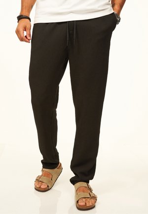 Man wearing black ribbed jogger pants with drawstring and beige buckle sandals, standing with hands in pockets.