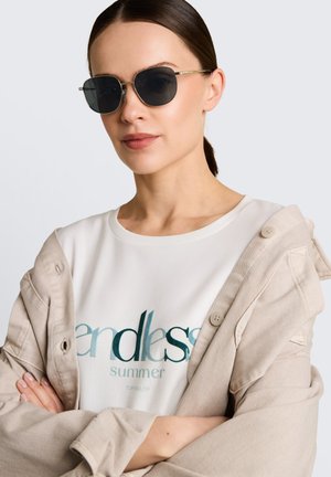 Femme portant des lunettes de soleil foncées, un t-shirt blanc "endless summer" et une veste beige posée sur ses épaules, les bras croisés.