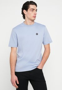 Svetlomodré bavlnené tričko s okrúhlym výstrihom, krátkymi rukávmi a malou čiernou nášivkou s logom na ľavej strane hrudníka. Prilieha k telu.