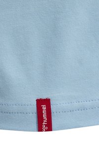 Lichtblauwe stof met een rood ingestikt label waarop witte "hummel" merktekst staat, vlak bij een gestikte zoom.