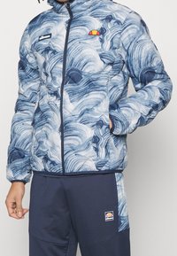 Person som bär en blå och vit jacka med virvlat mönster och marinblå byxor, båda med Ellesse-logotypen. Handen i jackfickan.