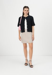 Zwarte gebreide cardigan met korte mouwen en knopen, witte aansluitende top, beige geribbelde rok, en zwarte high-heeled sandalen met stippenstrikken. Roze tas.