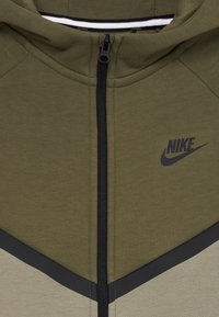 Olívazöld és világosszürke Nike zipzáras kapucnis pulóver, fekete cipzárral, kontrasztos háromszög mintás anyaggal és hímzett logóval.