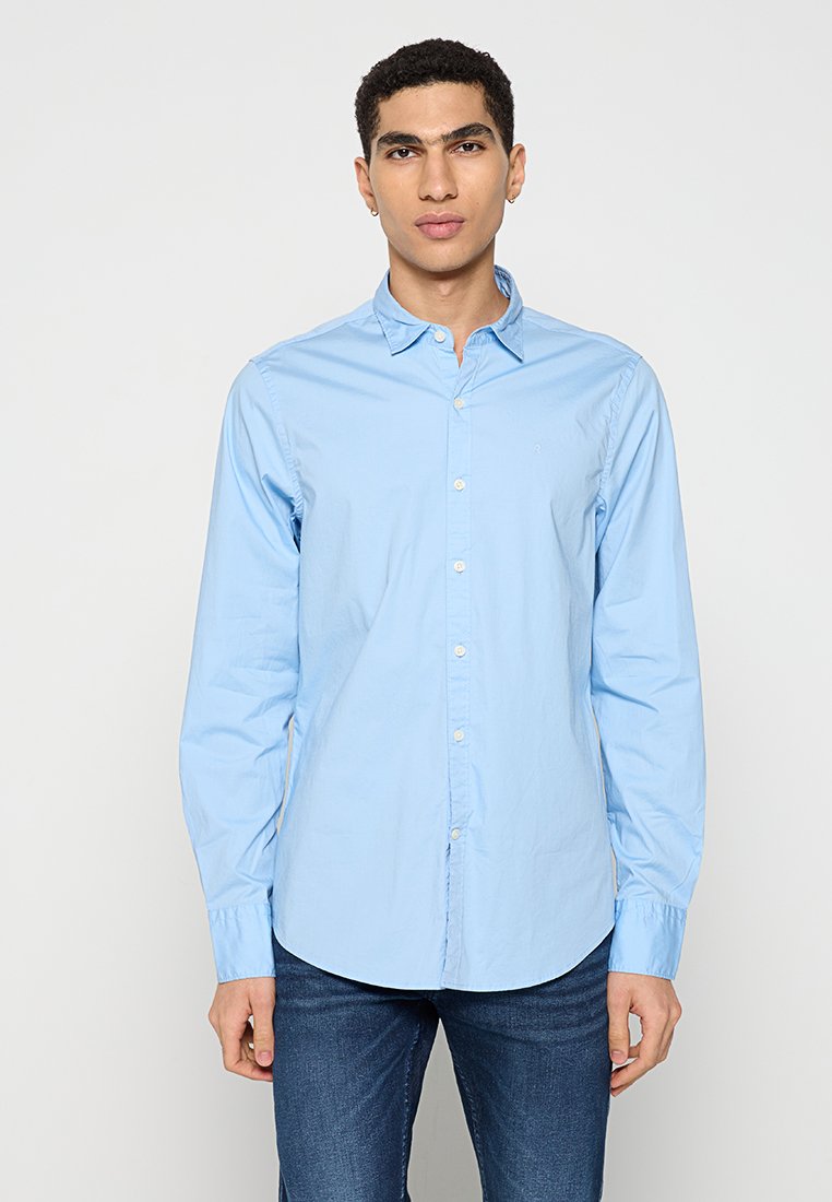 Camicetta azzurro chiaro con colletto, maniche lunghe e una tasca. Realizzata in un tessuto liscio; presenta bottoni sui polsini regolabili.