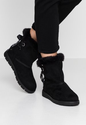 Bottes d'hiver noires avec doublure en fourrure, semelles épaisses et lacets décoratifs portées avec un pantalon noir sur fond blanc.
