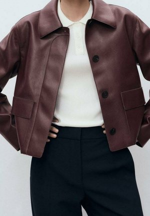 Personne portant une veste en cuir marron foncé sur une chemise blanche à col et un pantalon noir, mains reposées sur les hanches.