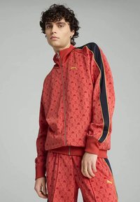 Roter Trainingsanzug mit einer Reißverschlussjacke und passenden Hosen. Der Stoff hat ein dezentes Muster mit marineblauen und gelben Akzenten an den Ärmeln.