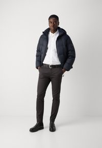 Veste en duvet marine avec capuche, fermeture éclair sur le devant, portée sur une chemise blanche, associée à un pantalon foncé et des bottines noires. Design simple et épuré.