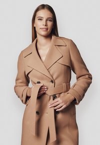 Beżowy wełniany płaszcz z podwójnym zapięciem, dużymi klapami i paskiem w talii. Posiada czarne guziki oraz detale na mankietach.