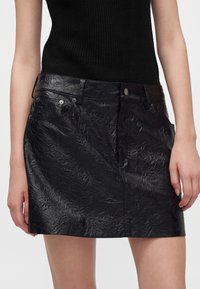 Femme portant une mini-jupe en cuir noir texturé et un haut en maille sans manches noir, montrée de la taille jusqu'au milieu de la cuisse.