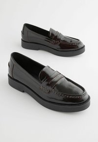 Mocassins en cuir verni noir avec une semelle en caoutchouc noire épaisse, présentant un cou-de-pied plissé et des détails de couture contrastants.