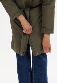 Olijfgroene waterdichte jas met een rits, klittenband manchetten en zijzakken over blauwe denim jeans. Voorzien van een eenvoudig ontwerp.