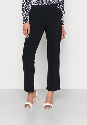 Woman wearing black straight-leg pants, patterned long-sleeve blouse, and white braided high-heeled sandals on a plain background.