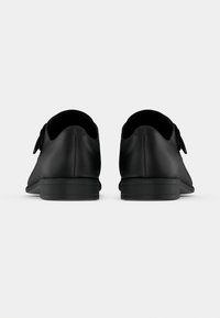 Μαύρα δερμάτινα παπούτσια με λεία υφή, στρογγυλευμένο σχήμα, χαμηλό τακούνι και ρυθμιζόμενο κούμπωμα με λουράκι. Πλάγια όψη που παρουσιάζει μινιμαλιστικό σχέδιο.