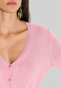Helle rosa Strickjacke mit V-Ausschnitt, die drei Knöpfe und eine weiche Textur aufweist. Große grüne tropfenförmige Ohrringe setzen einen Kontrast.