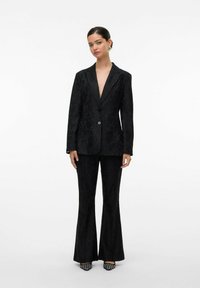 Blazer en velours noir et pantalon évasé avec un motif texturé. Présente un décolleté en V profond et une fermeture à un bouton, complété par des talons élégants.