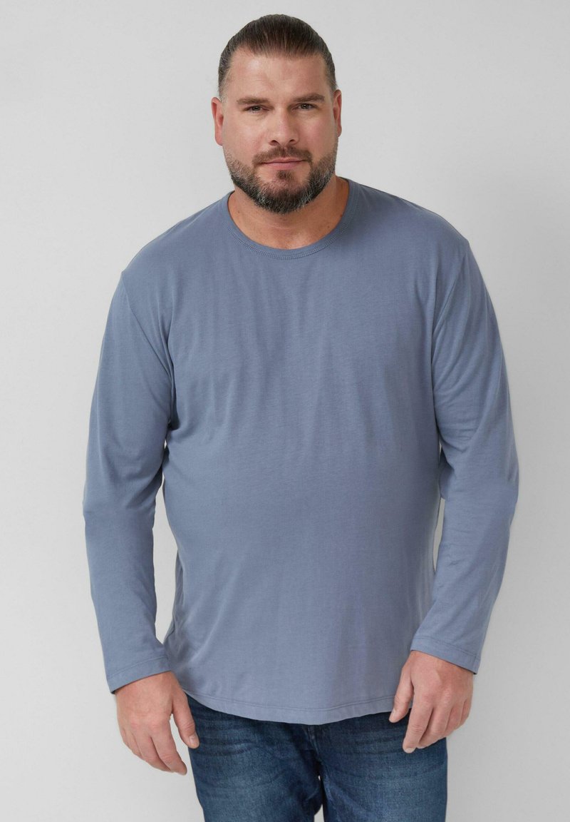 Langarmshirt aus weichem, hellblauem Stoff mit Rundhalsausschnitt und subtiler Drapierung, kombiniert mit blauen Jeans. Einfaches Design, ohne sichtbare Muster.