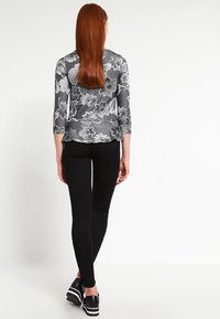 Woman with long red hair wearing a gray floral 3/4 sleeve top, black skinny pants, and black platform shoes walking away.