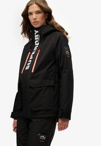 Czarna wodoodporna kurtka z wysokim kołnierzem, z jasnoróżowymi zamkami, wieloma kieszeniami oraz logo na kapturze i rękawie.