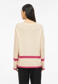 Femme aux longs cheveux bruns portant un pull beige avec des rayures horizontales roses à l'ourlet, aux manches et au dos, assorti à un pantalon noir, vue de dos.