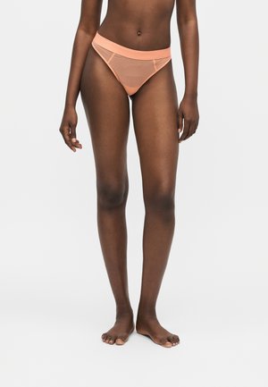 Femme debout pieds nus portant un sous-vêtement style bikini côtelé couleur pêche sur un fond clair uni.