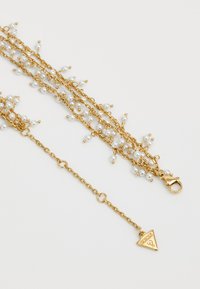 Gouden ketting met witte nepparels, met meerdere strengen en een kleine driehoekige hanger met "GUESS" gegraveerd. Gesp met kreeftsluitingen.