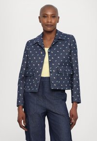 Donkerblauwe spijkerjack met bloemenpatroon, voorzien van een kraag, twee voorkruimelzakken en knoopsluitingen. Gestyled met een lichtgele top.