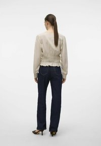 Beige satijnen blouse met elastische smocking op de taille, lange mouwen, gecombineerd met donkere rechte jeans en bruine haksandalen.