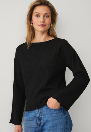 Kvinde med blondt hår iført guldøreringe, en struktureret sort langærmet top og blå jeans, med den ene hånd i lommen.