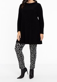 Schwarz langärmlige Tunika kombiniert mit leggings in schwarz-weißer Musterung, die giraffenartige Formen zeigen, ergänzt durch spitze schwarze Knöchelschäfte.