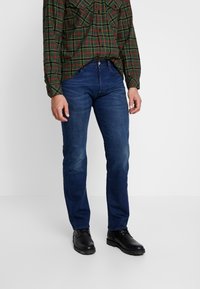 Jeans azul oscuro con un corte ajustado, detalles de desgaste ligero y diseño de cinco bolsillos, combinados con zapatos de cuero negros y una camisa de cuadros abotonada.
