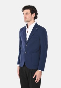 Blazer blu navy con design a un bottone, due tasche frontali e un sottile dettaglio di bottone. Indossato sopra una camicia bianca, con tessuto morbido.
