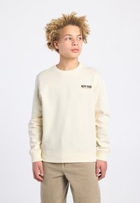 Sweatshirt de couleur crème avec un col rond, des manches longues et un petit logo "NEW YORK" sur la poitrine. Tissu doux et coupe décontractée.